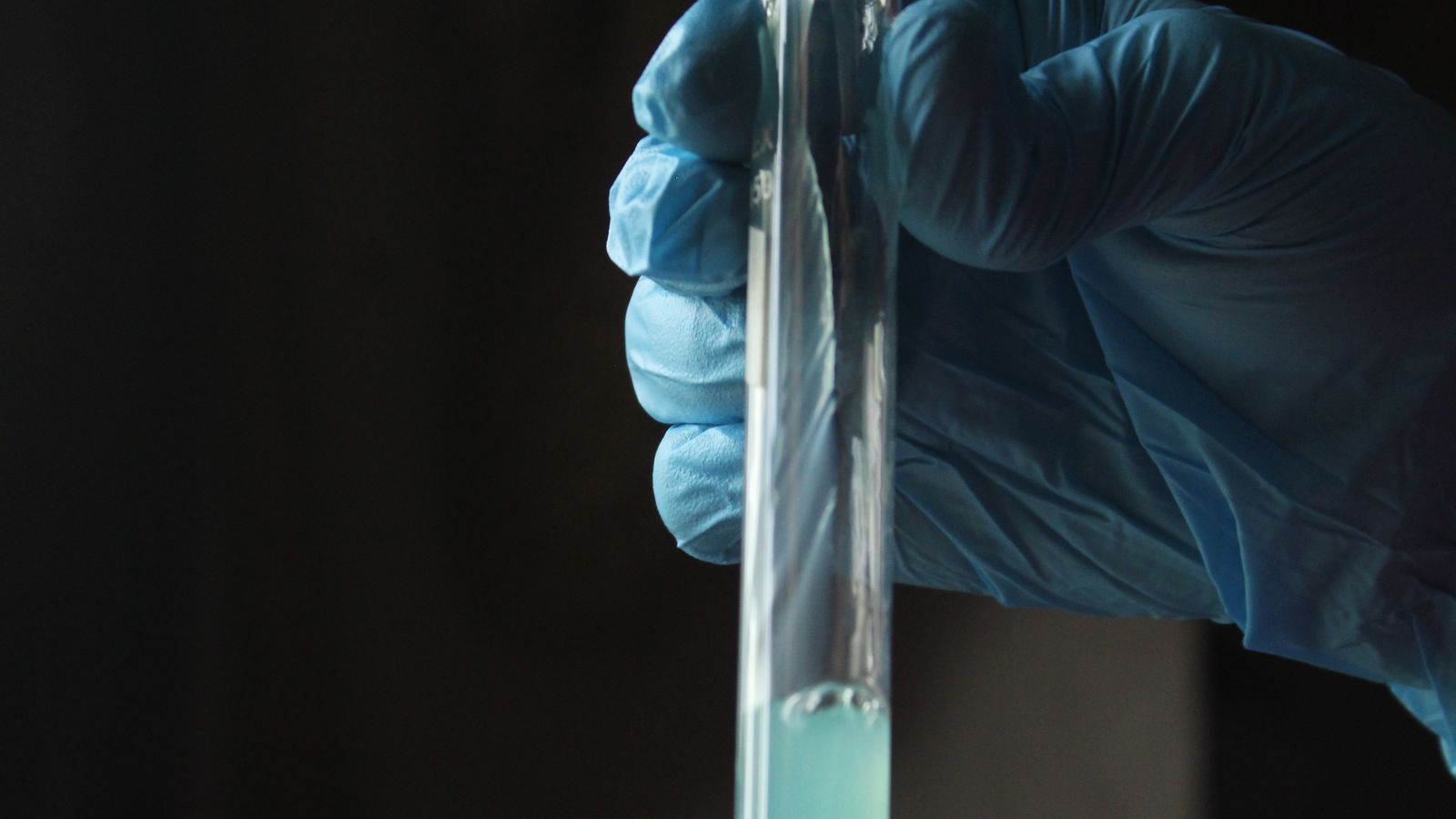 Gloved hands pouring a blue solution into a test tube in a laboratory setting.