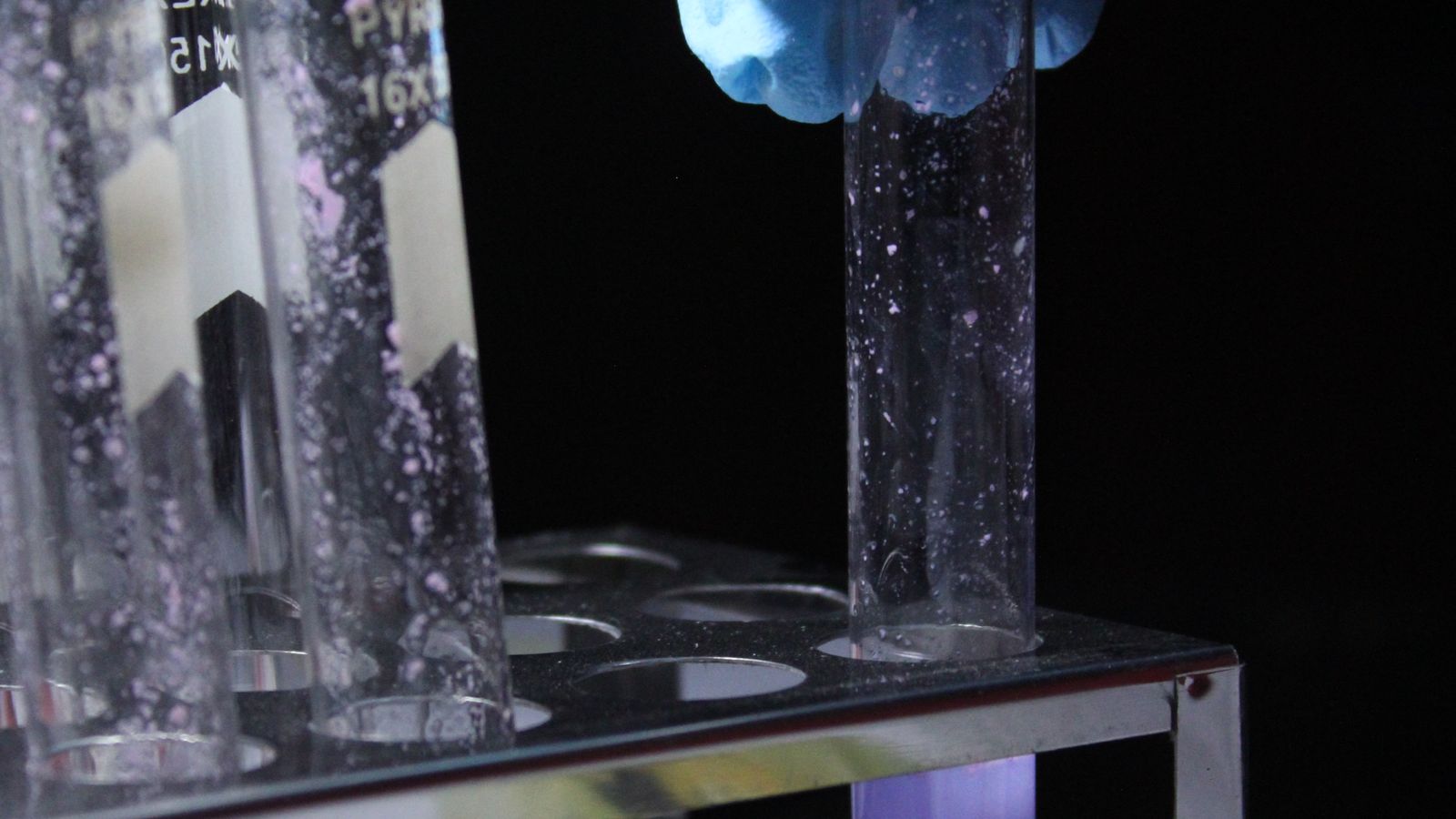 A gloved hand holds a small glass test tube containing a purple liquid in a laboratory setting.